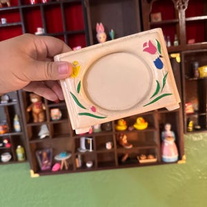 May include: A rectangular cream-colored ceramic frame with a central circular opening. The frame features hand-painted floral motifs including a yellow tulip, a pink tulip, and a blue tulip, each with green leaves, positioned around the center.