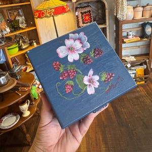 May include: A blue square box with a floral design featuring pink and white flowers and red berries. The box has a hand-painted look, with the artist's signature visible. The box is held in a hand.
