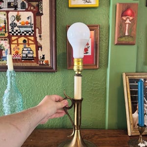 May include: A vintage brass candlestick lamp with a white bulb. The lamp has a cream-colored column and a decorative handle. The lamp is on a wooden surface, with framed artwork and a blue glass bottle in the background.