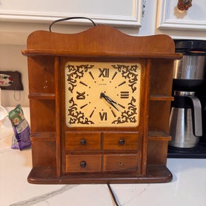 May include: A brown wooden wall clock featuring a square cream-colored face with black Roman numerals and ornate corner scrollwork. The clock includes three small shelves on each side, four small drawers below the face, and a power cord.