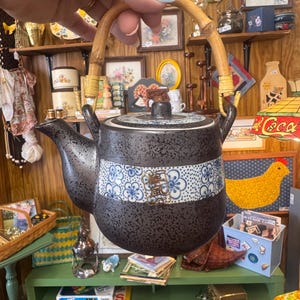 May include: A ceramic teapot with a dark gray speckled finish and a bamboo handle. The body features a central horizontal band decorated with a blue and white floral pattern and a gold-colored square emblem containing Japanese characters.