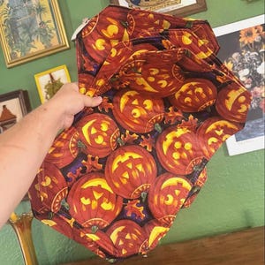 May include: A stack of Halloween-themed placemats featuring a repeating pattern of orange jack-o'-lanterns with yellow faces on a dark background. The placemats are made of fabric and have a quilted appearance. The overall color scheme is orange, yellow, and black.