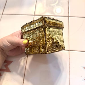May include: A square box covered in shimmering gold sequins with a matching lid. The box features dark brown trim along the edges and corners, providing a structured outline to the reflective, metallic surface of the small container.