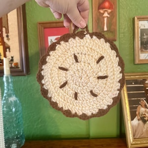 May include: A handmade crochet pie-shaped trivet with a light beige filling and a dark brown crust and decorative accents. The trivet has a loop for hanging and is held up by a hand.