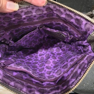 K&ouml;nnte beinhalten: Innenansicht einer braunen Umh&auml;ngetasche mit lila Leopardenmuster-Futter. Die Tasche hat mehrere F&auml;cher und einen Rei&szlig;verschluss. Der Innenraum ist leuchtend lila mit einem dunkleren Leopardenmuster.