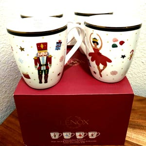 May include: Set of four white ceramic mugs with gold trim, featuring Nutcracker Ballet designs. Each mug showcases a different festive illustration, including a nutcracker soldier and a ballerina. The mugs are presented in a red box with the Lenox logo.