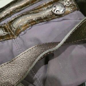 K&ouml;nnte beinhalten: Nahaufnahme einer metallisch silbernen Handtasche mit grauem Innenraum. Die Tasche verf&uuml;gt &uuml;ber mehrere Rei&szlig;verschl&uuml;sse, einen dekorativen silbernen Knopf und einen silbernen Riemen mit Schnalle. Das Design der Tasche deutet auf Stil und Funktionalit&auml;t hin.