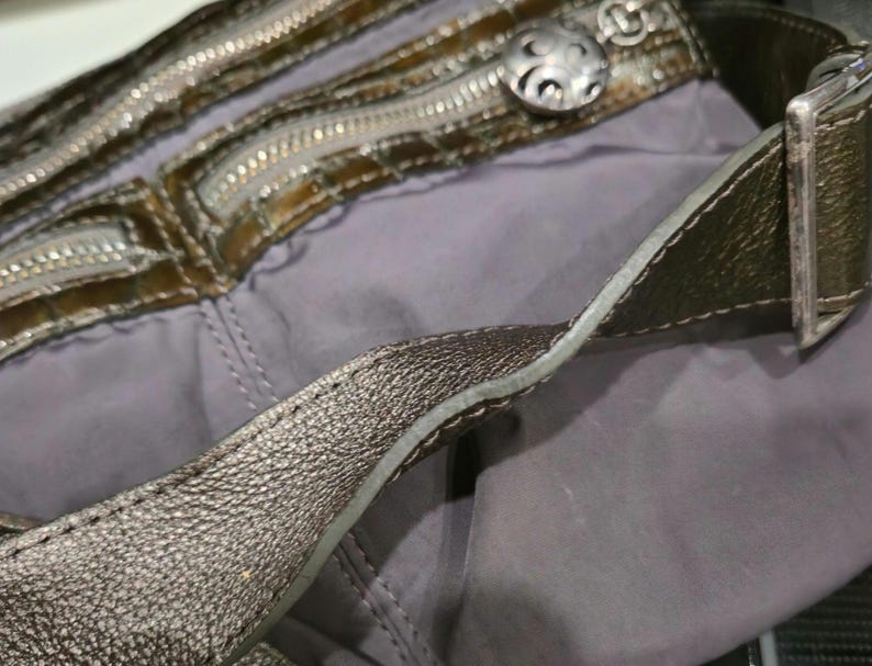 K&ouml;nnte beinhalten: Nahaufnahme einer metallisch silbernen Handtasche mit grauem Innenraum. Die Tasche verf&uuml;gt &uuml;ber mehrere Rei&szlig;verschl&uuml;sse, einen dekorativen silbernen Knopf und einen silbernen Riemen mit Schnalle. Das Design der Tasche deutet auf Stil und Funktionalit&auml;t hin.