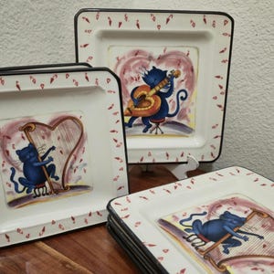May include: Set of square ceramic plates with a whimsical cat theme. Each plate features a blue cat playing a musical instrument, set against a pink heart background. The plates have a white base with a black border and red accents.