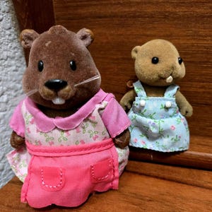 Op de afbeelding: Twee kleine, bruine beverbeeldjes. E&eacute;n draagt een roze jurk met een bloemenpatroon en een bijpassend schort. De andere draagt een blauwe bloemenoveralljurk. Beide hebben kleine, zwarte ogen en witte tanden.