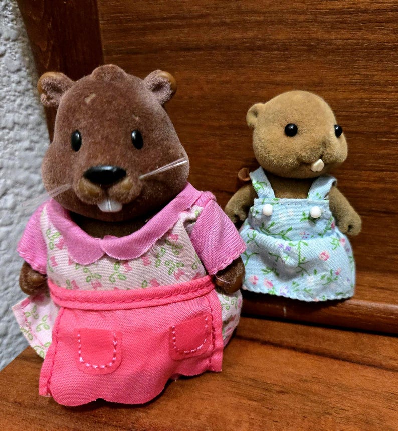 Op de afbeelding: Twee kleine, bruine beverbeeldjes. E&eacute;n draagt een roze jurk met een bloemenpatroon en een bijpassend schort. De andere draagt een blauwe bloemenoveralljurk. Beide hebben kleine, zwarte ogen en witte tanden.