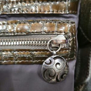 K&ouml;nnte beinhalten: Nahaufnahme einer braunen und grauen Handtasche mit einem strukturierten, krokodilartigen Muster. Die Tasche verf&uuml;gt &uuml;ber mehrere Rei&szlig;verschlussf&auml;cher mit silberfarbener Hardware und einen dekorativen, kreisf&ouml;rmigen Anh&auml;nger mit Wirbeldesign.