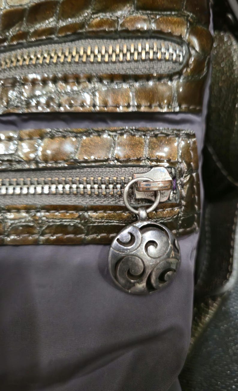 K&ouml;nnte beinhalten: Nahaufnahme einer braunen und grauen Handtasche mit einem strukturierten, krokodilartigen Muster. Die Tasche verf&uuml;gt &uuml;ber mehrere Rei&szlig;verschlussf&auml;cher mit silberfarbener Hardware und einen dekorativen, kreisf&ouml;rmigen Anh&auml;nger mit Wirbeldesign.