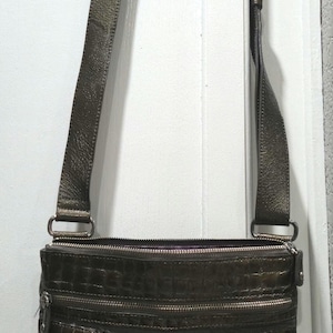 K&ouml;nnte beinhalten: Dunkelbraune Umh&auml;ngetasche mit einem langen, verstellbaren Riemen. Die Tasche verf&uuml;gt &uuml;ber mehrere Rei&szlig;verschlussf&auml;cher und dekorative silberfarbene Beschl&auml;ge. Der untere Teil der Tasche ist dunkler.