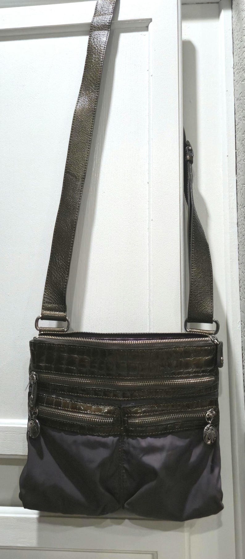 K&ouml;nnte beinhalten: Dunkelbraune Umh&auml;ngetasche mit einem langen, verstellbaren Riemen. Die Tasche verf&uuml;gt &uuml;ber mehrere Rei&szlig;verschlussf&auml;cher und dekorative silberfarbene Beschl&auml;ge. Der untere Teil der Tasche ist dunkler.