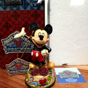 May include: A Mickey Mouse figurine from the Disney Traditions collection. The figure is black, red, and beige, standing on a decorative base with roses. The figurine is part of the Walt Disney Showcase Collection.