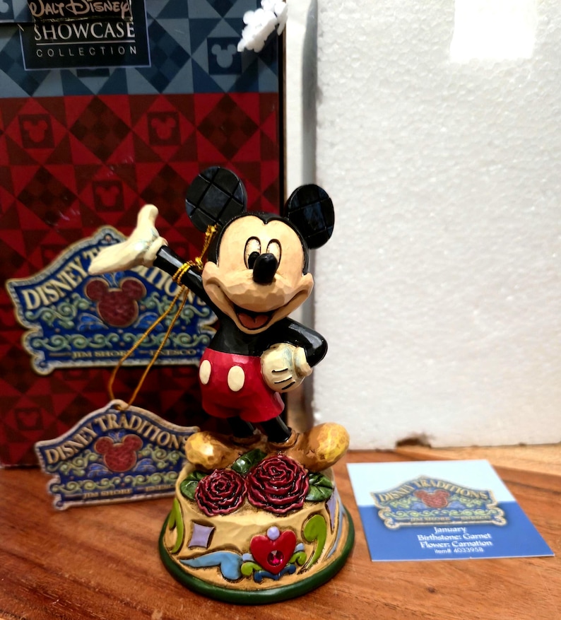 May include: A Mickey Mouse figurine from the Disney Traditions collection. The figure is black, red, and beige, standing on a decorative base with roses. The figurine is part of the Walt Disney Showcase Collection.