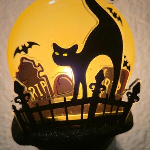 May include: A Halloween-themed night light featuring a black cat silhouette on a graveyard scene with a yellow moon backdrop. The cat stands on a fence in front of tombstones, with bats flying around the moon. The night light has a black base.