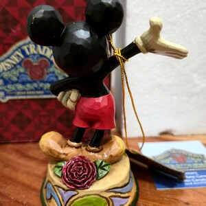 May include: A Mickey Mouse figurine with black ears, a black body, red shorts, and tan shoes. The figurine is standing on a decorative base with a red rose and green leaves. A gold string is attached to the figurine.