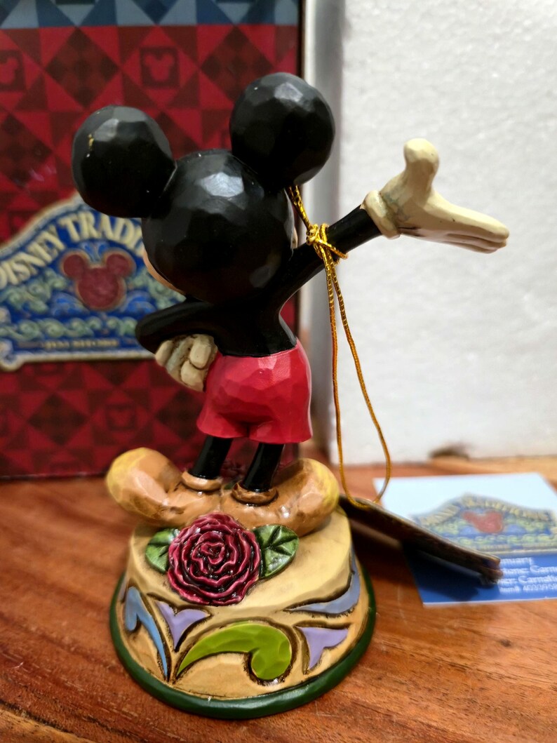 May include: A Mickey Mouse figurine with black ears, a black body, red shorts, and tan shoes. The figurine is standing on a decorative base with a red rose and green leaves. A gold string is attached to the figurine.