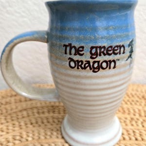 May include: A ceramic mug with a flared rim and a pedestal base, featuring a ribbed texture. The upper portion is glazed in blue, transitioning to a cream color on the lower body. The text The Green Dragon is printed in dark brown lettering.