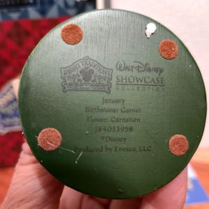 May include: A green, circular Disney Traditions collectible base with text including "January", "Birthstone: Garnet", and "Flower: Carnation". The base has four brown felt pads and the text "Walt Disney Showcase Collection".