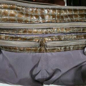 K&ouml;nnte beinhalten: Eine braune und graue Umh&auml;ngetasche mit mehreren Rei&szlig;verschlussf&auml;chern. Der obere Bereich hat ein strukturiertes, krokodilartiges Muster, w&auml;hrend der untere Bereich aus glattem, grauem Material besteht. Silberfarbene Hardware und dekorative Anh&auml;nger.