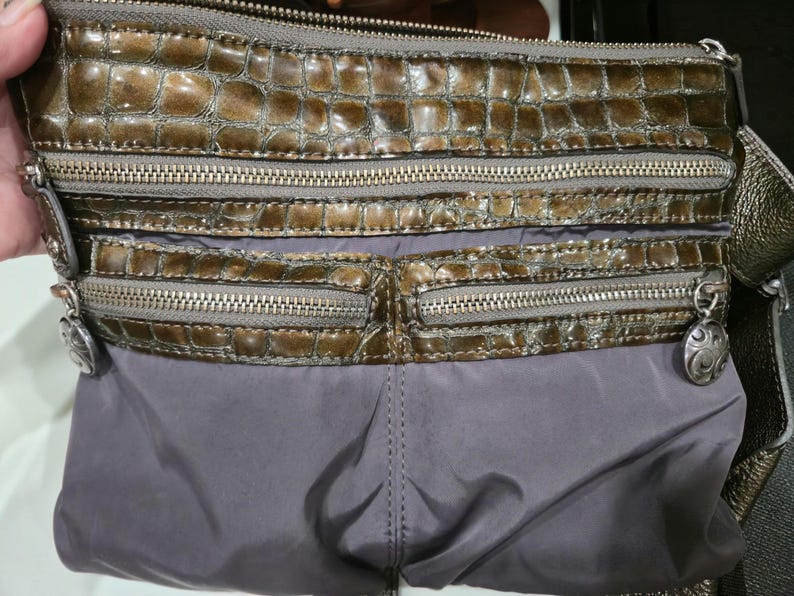 K&ouml;nnte beinhalten: Eine braune und graue Umh&auml;ngetasche mit mehreren Rei&szlig;verschlussf&auml;chern. Der obere Bereich hat ein strukturiertes, krokodilartiges Muster, w&auml;hrend der untere Bereich aus glattem, grauem Material besteht. Silberfarbene Hardware und dekorative Anh&auml;nger.