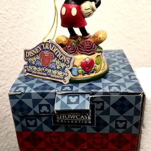 May include: A Disney Traditions figurine of Mickey Mouse in a classic pose, standing on a decorative base with roses and a heart. The figurine is atop a box with the Disney Traditions logo.