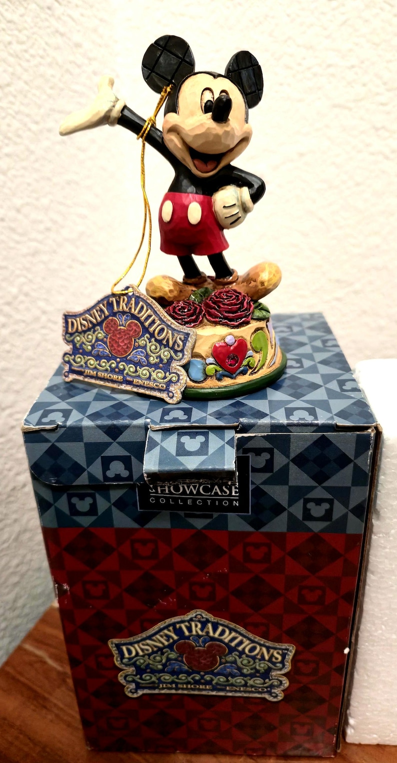 May include: A Disney Traditions figurine of Mickey Mouse in a classic pose, standing on a decorative base with roses and a heart. The figurine is atop a box with the Disney Traditions logo.