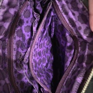 K&ouml;nnte beinhalten: Innenansicht einer schwarzen Handtasche mit lila Leopardenmuster-Futter. Die Tasche hat mehrere Rei&szlig;verschlussf&auml;cher und einen dunkelvioletten Rei&szlig;verschluss. Das Futter ist ein leuchtendes Lila mit einem schwarzen Leopardenmuster.
