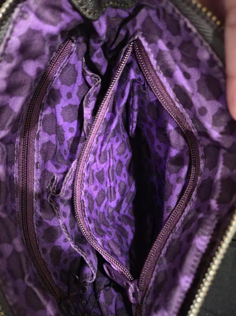 K&ouml;nnte beinhalten: Innenansicht einer schwarzen Handtasche mit lila Leopardenmuster-Futter. Die Tasche hat mehrere Rei&szlig;verschlussf&auml;cher und einen dunkelvioletten Rei&szlig;verschluss. Das Futter ist ein leuchtendes Lila mit einem schwarzen Leopardenmuster.