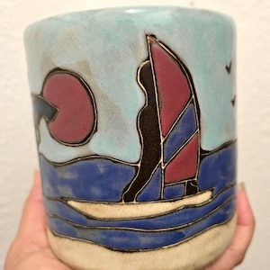 May include: A cylindrical ceramic mug featuring a hand-painted scene of a sailboat with a maroon and blue sail on dark blue water. The design includes a large maroon sun in a light blue sky, a sandy beige base, and black outlines defining the shapes.