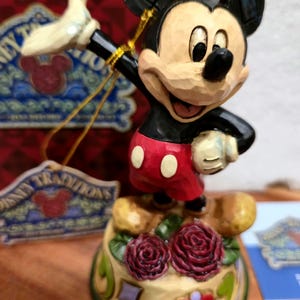 May include: A Mickey Mouse figurine in a classic pose, wearing red shorts with white buttons, a black jacket, and yellow gloves. The figurine stands on a base decorated with roses and hearts. The figurine is part of the Disney Traditions collection.