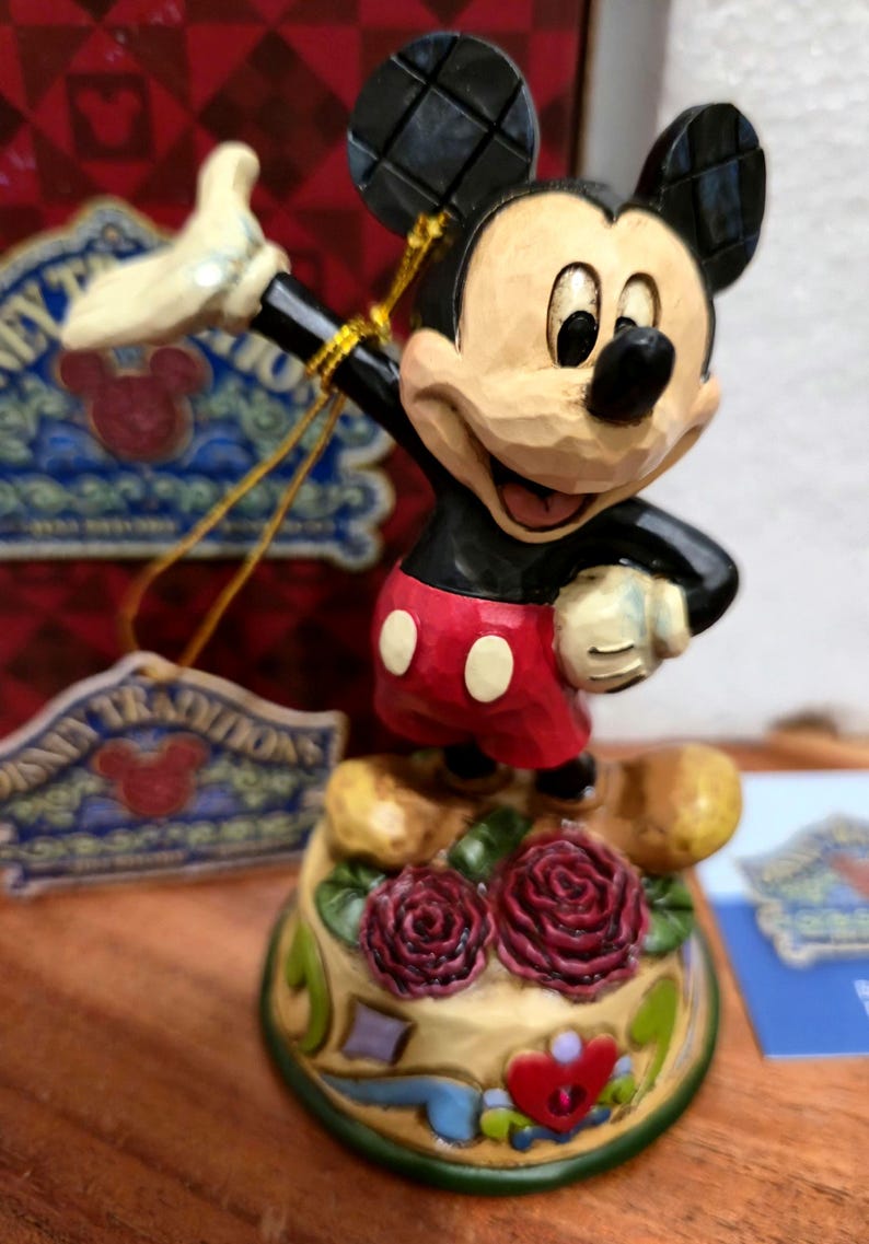 May include: A Mickey Mouse figurine in a classic pose, wearing red shorts with white buttons, a black jacket, and yellow gloves. The figurine stands on a base decorated with roses and hearts. The figurine is part of the Disney Traditions collection.