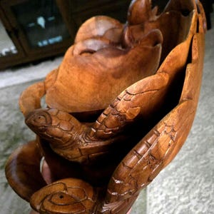 May include: A brown wooden bowl featuring carved sea turtle figures integrated into the exterior rim. The bowl has a smooth, polished finish with visible wood grain patterns and detailed etchings on the turtle shells and heads.
