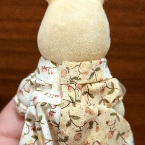 Op de afbeelding: Een kleine, cr&egrave;mekleurige konijnenfiguur met een outfit met bloemenprint. De outfit heeft een witte achtergrond met roze en groene bloempatronen. De oren van het konijn staan rechtop en het algehele ontwerp is zacht en grillig.