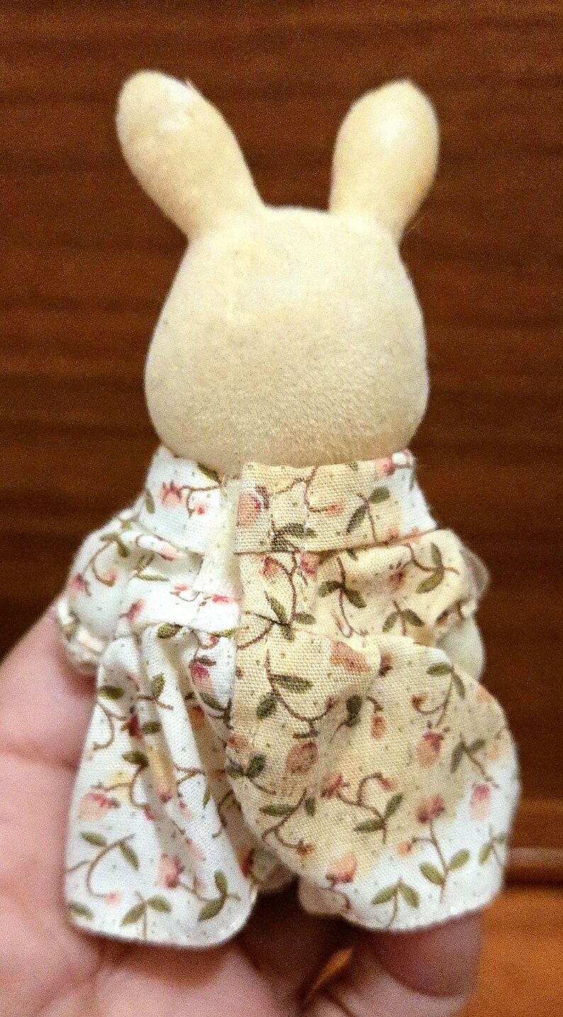 Op de afbeelding: Een kleine, cr&egrave;mekleurige konijnenfiguur met een outfit met bloemenprint. De outfit heeft een witte achtergrond met roze en groene bloempatronen. De oren van het konijn staan rechtop en het algehele ontwerp is zacht en grillig.