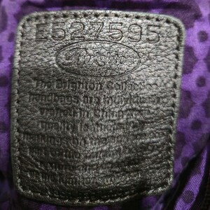 K&ouml;nnte beinhalten: Nahaufnahme eines schwarzen Lederetiketts mit dem gepr&auml;gten Text "E527595" und dem Brighton-Logo. Das Etikett ist auf einem lila Stoff mit Rautenmuster befestigt. Das Etikett enth&auml;lt auch Text &uuml;ber die Handtaschen der Brighton Collection.