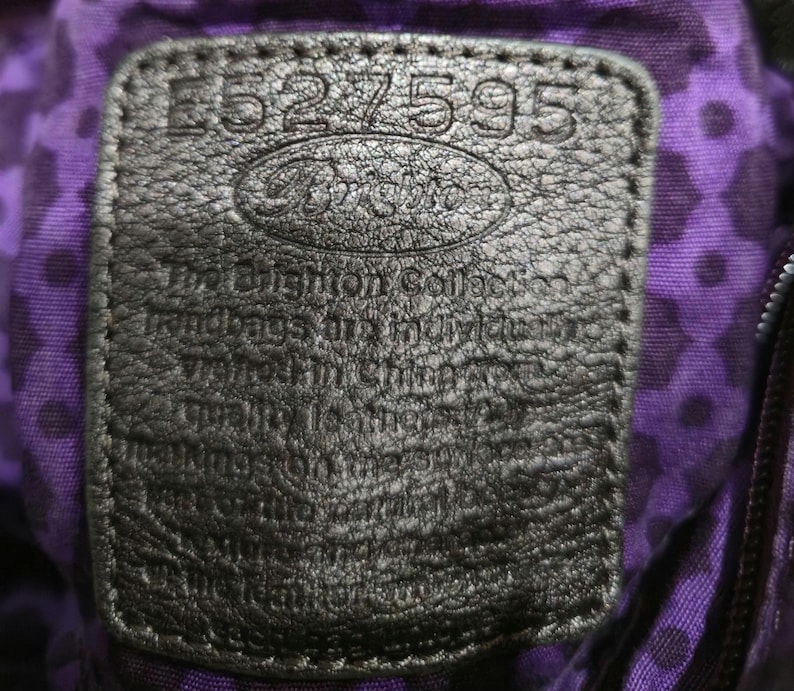 K&ouml;nnte beinhalten: Nahaufnahme eines schwarzen Lederetiketts mit dem gepr&auml;gten Text "E527595" und dem Brighton-Logo. Das Etikett ist auf einem lila Stoff mit Rautenmuster befestigt. Das Etikett enth&auml;lt auch Text &uuml;ber die Handtaschen der Brighton Collection.