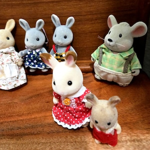 Op de afbeelding: Een verzameling miniatuur dierfiguren, waaronder konijnen en een muis, gekleed in verschillende outfits. De konijnen dragen jurken en overalls, terwijl de muis een groen geruit overhemd met bretels draagt. De figuren staan op een houten oppervlak.