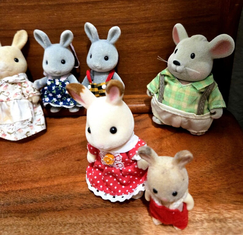 Op de afbeelding: Een verzameling miniatuur dierfiguren, waaronder konijnen en een muis, gekleed in verschillende outfits. De konijnen dragen jurken en overalls, terwijl de muis een groen geruit overhemd met bretels draagt. De figuren staan op een houten oppervlak.