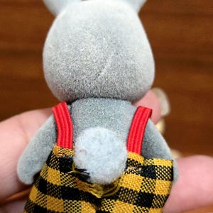 Op de afbeelding: Een grijze konijnenfiguur met een geel en zwart geruite overall met rode bretels. Het konijn heeft een pluizige staart en rechtopstaande oren. De figuur is een speelgoed.