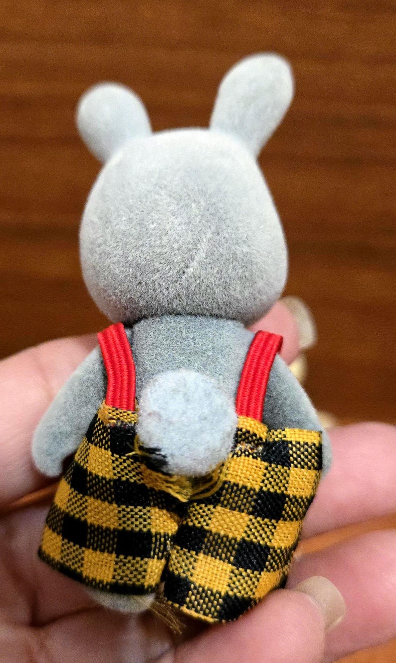 Op de afbeelding: Een grijze konijnenfiguur met een geel en zwart geruite overall met rode bretels. Het konijn heeft een pluizige staart en rechtopstaande oren. De figuur is een speelgoed.
