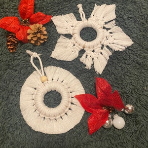 May include: A collection of handmade Christmas ornaments and decorations. The ornaments include a white macrame snowflake, a circular macrame ornament, and red poinsettia-style decorations with pinecones and silver and white baubles. The items are arranged on a dark green surface.