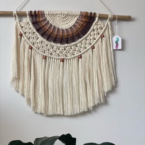 May include: A cream-colored macrame wall hanging with a half-circle design. The top section features a woven pattern with brown and tan accents. The bottom has long, flowing fringe. Wooden beads are evenly spaced along the bottom edge. The wall hanging is suspended from a wooden dowel.