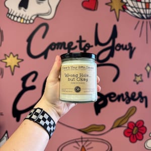 May include: A clear glass candle jar with a black lid and a beige label. The label reads: Come to Your Effin Senses, Wrong Hole... but Okay, fudge nut brownie, 8 oz (226 g), soy wax and fragrance. The candle contains white wax.