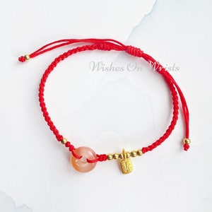 May include: Red string bracelet with a gold charm and a peach-colored stone. The charm has Chinese characters on it. The bracelet is tied with a knot and has gold beads.