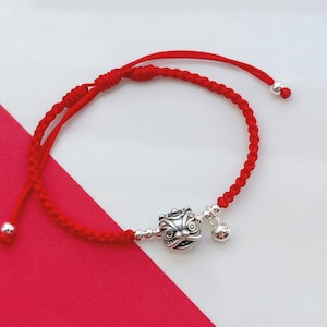 May include: Red string bracelet with a silver lion dance head charm and a silver bell charm.