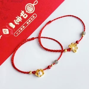 May include: Two red string bracelets with gold flower charms and pearl centres. The bracelets also feature small gold beads and grey beads. The background includes a red card with gold Chinese characters.
